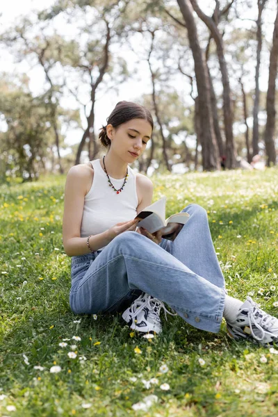 Jeune femme lisant le livre assis sur la prairie avec des fleurs dans le parc — Photo de stock