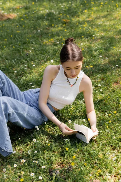 Вид под высоким углом на молодую женщину в повседневной одежде, читающую книгу о мясорубке с цветами в парке — стоковое фото