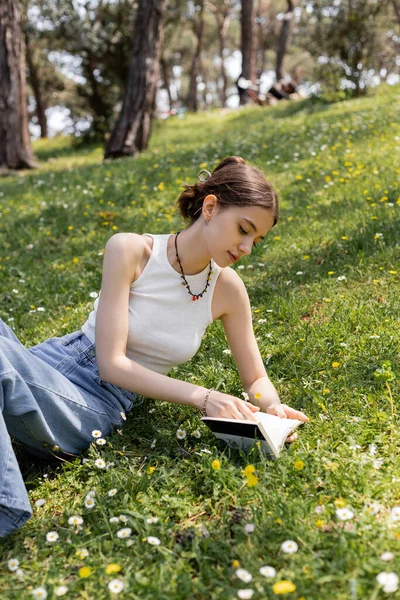 Young woman in top reading book while lying on lawn with flowers in park — Stock Photo