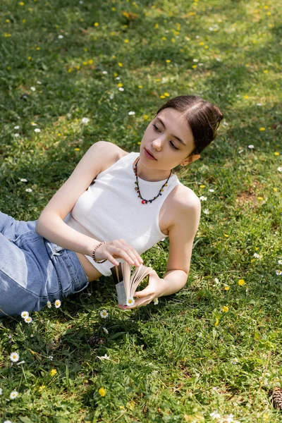 High angle view of young brunette woman in top and jeans holding book while lying on lawn with daisies in park — Stock Photo