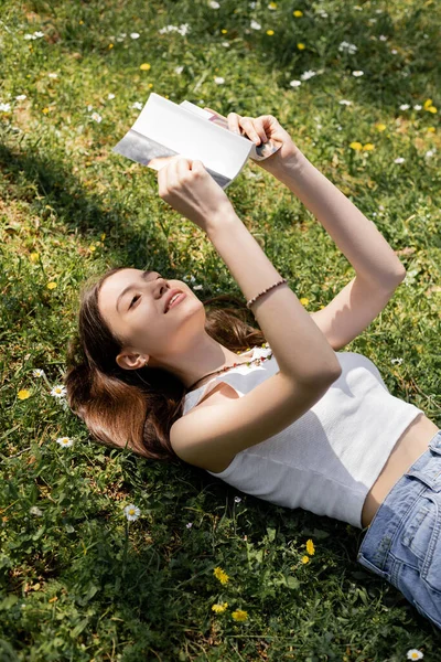 High angle view of smiling young woman reading book while lying on meadow with flowers in park — Stock Photo
