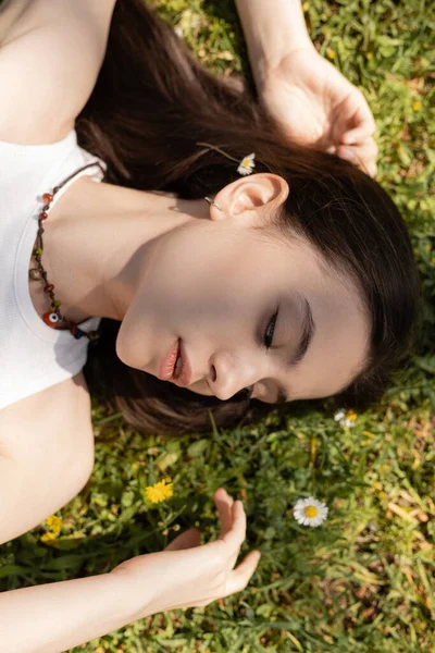 Visão superior da jovem morena olhando para flores enquanto estava deitada no gramado no parque — Fotografia de Stock