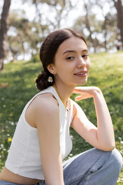Портрет улыбающейся молодой женщины с цветком в волосах, смотрящей в камеру в летнем парке — стоковое фото