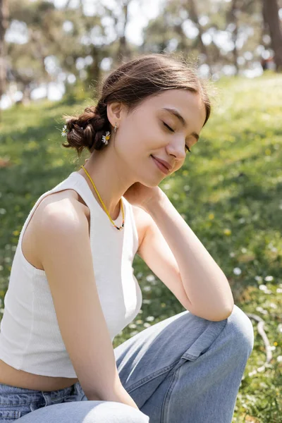 Joyeux jeune femme avec des fleurs dans les cheveux assis sur la pelouse floue dans le parc d'été — Photo de stock