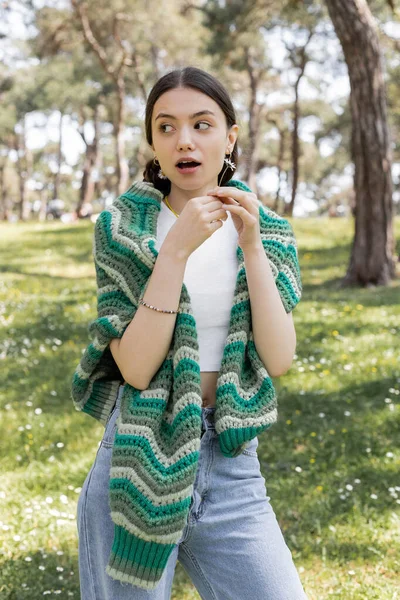 Surprised woman with knitted sweater on shoulders holding daisy in summer park — Stock Photo