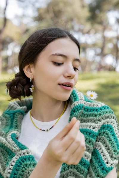 Giovane donna bruna con maglione lavorato a maglia sulle spalle tenendo sfocato fiore di margherita nel parco — Foto stock