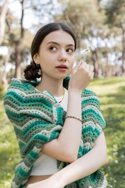 Portrait de jeune femme avec pull tricoté sur les épaules tenant marguerite dans le parc — Photo de stock