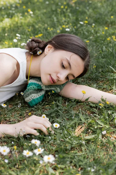 Retrato de jovem morena olhando para margaridas borradas enquanto deitado no prado no parque — Fotografia de Stock
