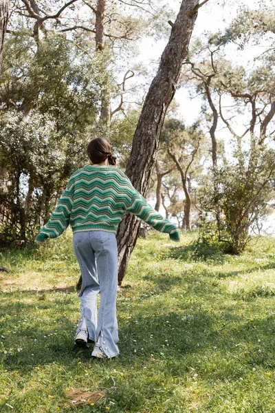Vista posteriore di giovane donna in jeans e maglione che cammina nel parco estivo — Foto stock