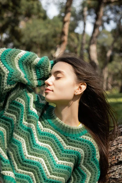 Young woman in knitted sweater touching hair near tree in park — Stock Photo