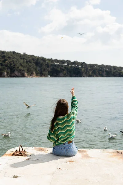 Back view of young woman sitting on pier near blurred seagulls on water in Turkey — Stock Photo