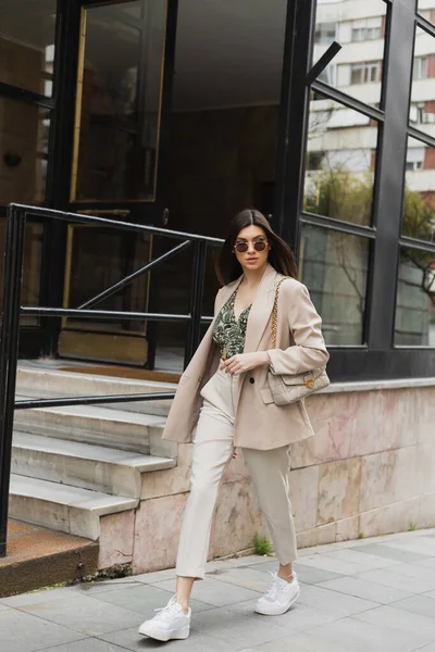 Jeune femme dans des lunettes de soleil élégantes et tenue à la mode avec pantalon blanc et blazer beige marche avec sac à main près du bâtiment moderne avec des fenêtres et des escaliers dans la rue à Istanbul — Photo de stock
