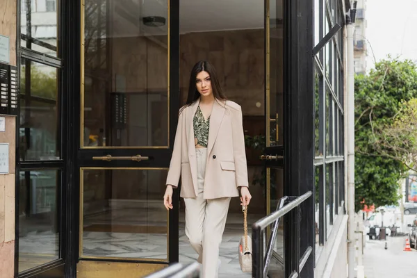 Brunette and young woman in trendy outfit with white pants, blouse and beige blazer walking out of modern building while holding handbag with chain strap on street in Istanbul — Stock Photo
