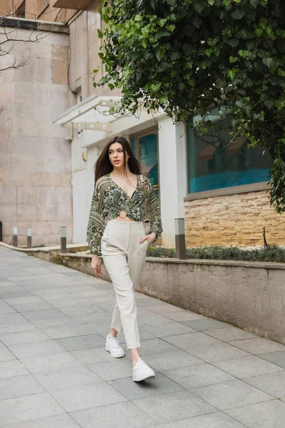 Модная женщина с длинными волосами в модном наряде с бежевыми брюками, обрезанной блузкой и сумочкой на цепном ремне, идущая с рукой в кармане рядом с современным зданием и зеленым деревом на улице в Стамбуле — стоковое фото