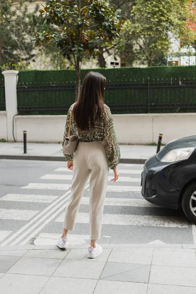 На задньому плані - шикарна жінка з довгим волоссям в модному одязі з бежевими штанями, затиснутою блузкою і сумочкою з ланцюговим ременем, що стоїть на перехресті біля машини на міській вулиці в Стамбулі. — стокове фото