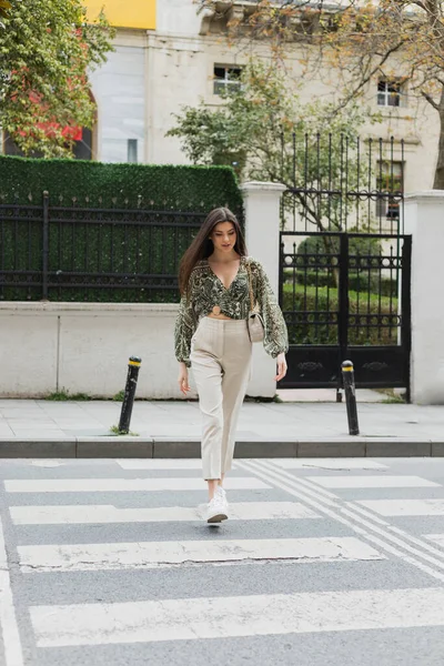 Mujer elegante con pelo largo morena en traje de moda con pantalones beige, blusa recortada y bolso con correa de cadena caminando en el paso de calle urbana en Estambul, casa borrosa en el fondo - foto de stock