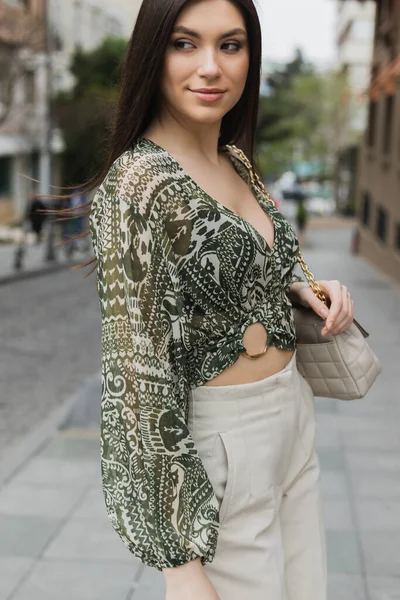 Femme élégante avec brune cheveux longs en tenue tendance avec pantalon beige, chemisier recadré et sac à main avec sangle de chaîne debout et souriant dans la rue urbaine d'Istanbul — Photo de stock