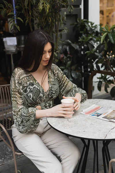 Молодая женщина с длинными брюнетками и макияжем сидит на стуле возле круглого стола бистро с газетой и держит бумажную чашку с кофе возле размытых растений на террасе кафе в Стамбуле — стоковое фото