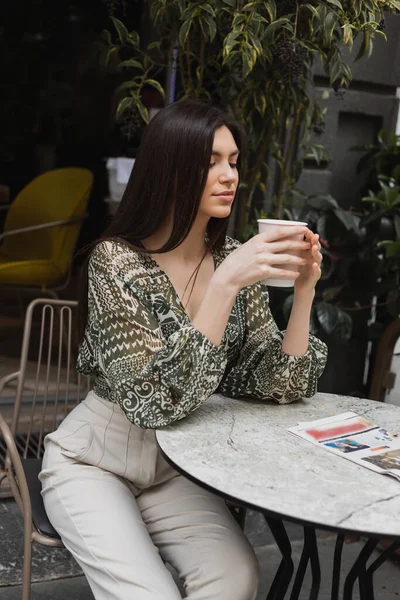 Séduisante femme aux longs cheveux bruns et maquillage assis sur une chaise près d'une table de bistrot ronde avec journal et tenant une tasse en papier avec café près de plantes floues sur la terrasse du café à Istanbul — Photo de stock