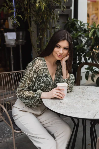 Dreamy woman with long brunette hair and makeup sitting on chair near round bistro table and holding paper cup with coffee near blurred plants on terrace of cafe in Istanbul — Stock Photo