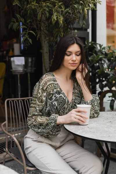 Young woman with long brunette hair and makeup sitting on chair near round bistro table and looking at coffee in paper cup near blurred plants on terrace of cafe in Istanbul — Stock Photo