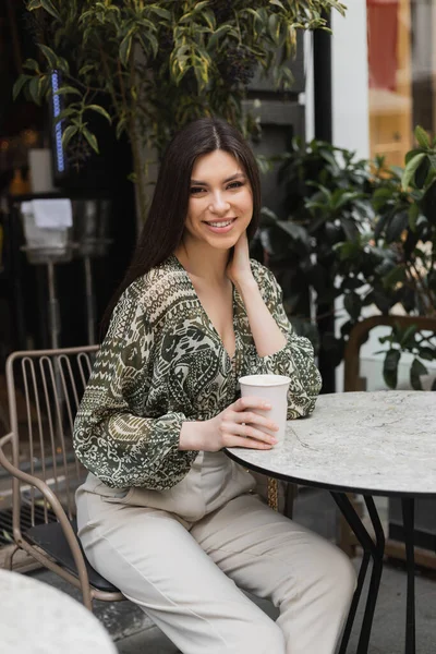 Радісна молода жінка з довгим брюнетним волоссям і гримом сидить на стільці біля круглого бістро столу і тримає каву в паперовій чашці і посміхається біля розмитих рослин на терасі кафе в Стамбулі. — стокове фото