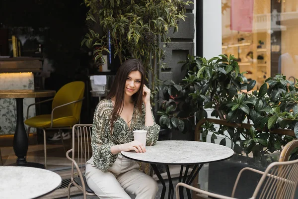 Smiling woman with long brunette hair and makeup sitting on chair near round bistro table and holding coffee in paper cup near blurred plants on terrace of cafe in Istanbul — Stock Photo