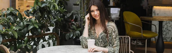 Smiling young woman with long brunette hair sitting on chair near round bistro table and holding coffee in paper cup near blurred plants on terrace of cafe in Istanbul, banner — Stock Photo