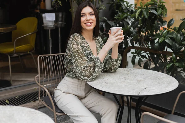 Очаровательная женщина с длинными волосами сидит на стуле возле круглого стола бистро и держит бумажный стакан с напитком, глядя в сторону и улыбаясь рядом размытые растения на террасе кафе в Стамбуле — стоковое фото
