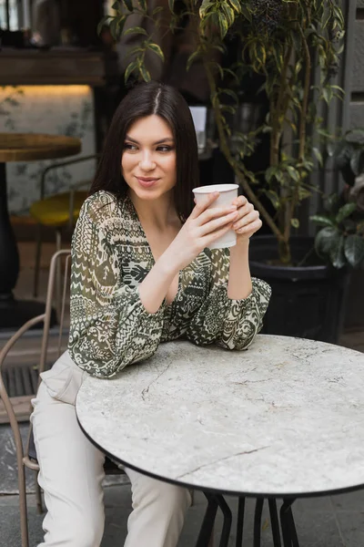 Young woman with long brunette hair sitting on chair near round bistro table and holding paper cup with drink while looking away and smiling near blurred plants on terrace of cafe in Istanbul — Stock Photo