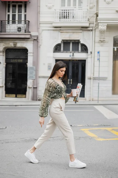 Bonita joven con pelo largo morena y maquillaje sosteniendo taza de papel con café y periódico mientras camina en traje de moda con pantalones beige y blusa en la calle urbana cerca del edificio en Estambul - foto de stock