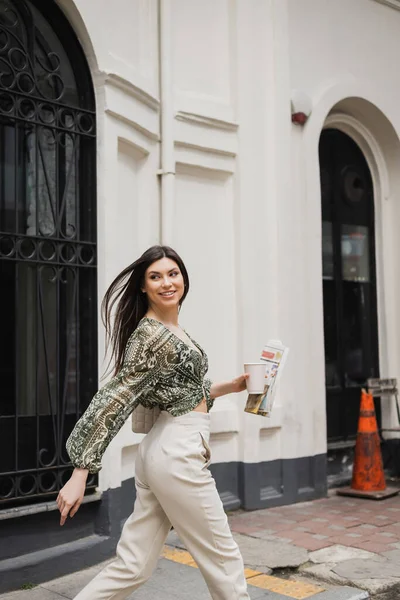 Positive woman with long brunette hair and makeup holding paper cup with coffee and newspaper while walking in trendy outfit and handbag on urban street near metallic fence and white wall in Istanbul — Stock Photo