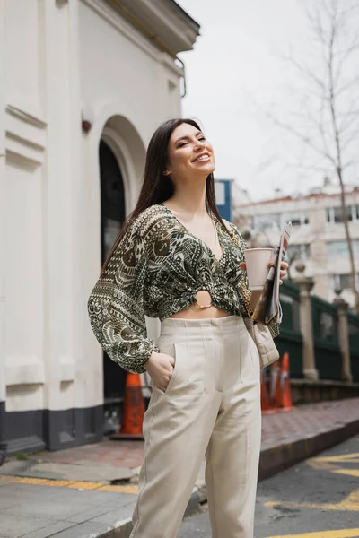 Позитивна жінка з довгим волоссям і гримом, що тримає паперову чашку з кавою і газетою, стоячи в модному спорядженні з сумочкою на міській вулиці біля білого будинку в Стамбулі. — стокове фото