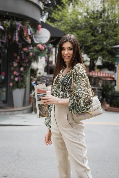 Chic woman with long hair holding paper cup with coffee and newspaper while walking in trendy outfit with handbag on chain strap and smiling on urban street near blurred flower shop in Istanbul — Stock Photo