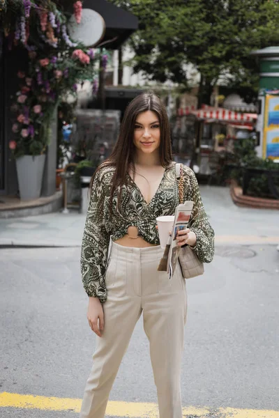 Bela mulher com cabelo longo segurando copo de papel com café e jornal, enquanto em pé na roupa da moda com bolsa na cadeia cinta e sorrindo na rua urbana perto de loja de flores borradas em Istambul — Fotografia de Stock