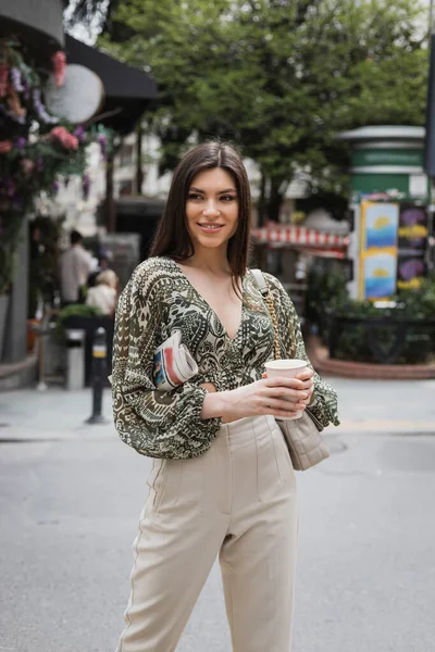 Beautiful woman with long hair holding morning coffee in paper cup and newspaper while standing in trendy outfit with handbag and smiling on urban street near blurred flower shop in Istanbul — Stock Photo