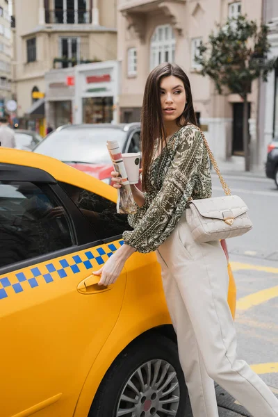 Chic woman with long hair holding paper cup with coffee and newspaper while standing in trendy outfit with handbag on chain strap and opening door of yellow taxi on blurred urban street in Istanbul — Stock Photo