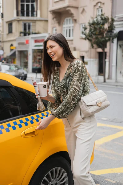 Femme heureuse avec les cheveux longs tenant tasse en papier avec café et journal tout en se tenant dans une tenue à la mode avec sac à main et ouvrir la porte de taxi jaune sur la rue urbaine floue à Istanbul — Photo de stock