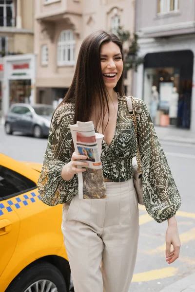 Mujer positiva con cabello largo sosteniendo taza de papel con café y periódico mientras camina en traje de moda con bolso en correa de cadena cerca de taxi amarillo en la calle urbana borrosa en Estambul - foto de stock