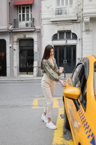 Шикарна жінка з довгим волоссям тримає каву в паперовій чашці, стоячи в модному вбранні з сумочкою на ланцюговому ремені і відкриваючи двері жовтого таксі на розмитій міській вулиці — стокове фото