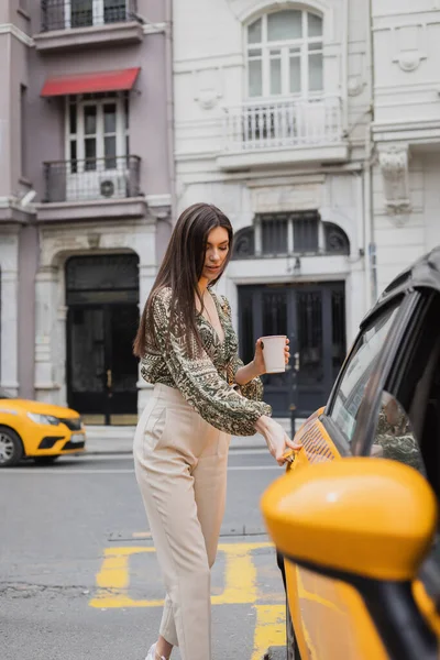 Mulher atraente com cabelos longos segurando café em copo de papel enquanto estava de pé na roupa da moda com bolsa e porta de abertura da cabine amarela na rua urbana turva com edifício em Istambul — Fotografia de Stock