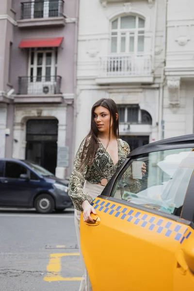 Стильная женщина с длинными волосами, держащая кофе в бумажной чашке, стоя в модном наряде с сумочкой на цепном ремне и открывая дверь желтого такси, глядя на размытую улицу Стамбула — стоковое фото