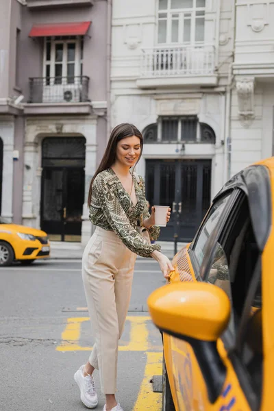 Lächelnde junge Frau mit langen Haaren, die Kaffee in Pappbecher hält, während sie im trendigen Outfit mit Handtasche am Kettenriemen steht und die Tür des gelben Taxis auf der verschwommenen Stadtstraße in Istanbul öffnet — Stockfoto