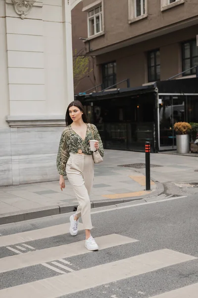 Стильная женщина с длинными волосами в модном наряде с бежевыми брюками, обрезанной блузкой и сумочкой с цепным ремнем идущей по пешеходной дорожке с кофе на городской улице с размытым зданием в Стамбуле — стоковое фото