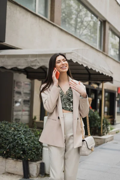 Smiling young woman with brunette long hair and makeup smiling while talking on smartphone and standing in trendy outfit with handbag on chain strap near blurred restaurant in Istanbul — Stock Photo