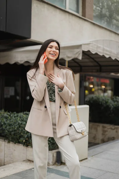 Hermosa mujer con cabello largo morena y maquillaje sonriendo mientras habla en el teléfono inteligente y de pie en traje de moda con bolso de mano en correa de cadena cerca de un restaurante de lujo borroso en Estambul - foto de stock