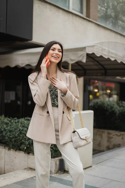 Belle femme aux cheveux longs brunes et au maquillage souriant tout en parlant sur smartphone et debout en tenue tendance avec sac à main sur sangle chaîne près d'un restaurant chic flou à Istanbul — Photo de stock