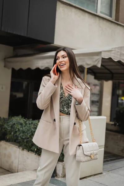 Позитивная молодая женщина с брюнеткой длинные волосы и макияж улыбаясь во время разговора на смартфоне и стоя в модном наряде с сумочкой на ремне цепи рядом размытый модный ресторан в Стамбуле — стоковое фото