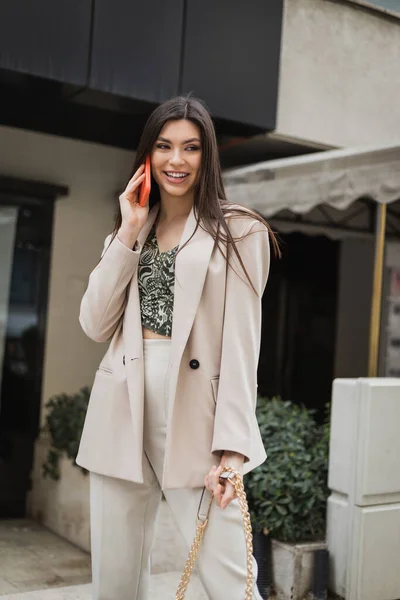 Счастливая женщина с длинными волосами и макияжем улыбается, разговаривая по смартфону и стоя в модном наряде с сумочкой на ремне рядом с размытым модным рестораном в Стамбуле — стоковое фото