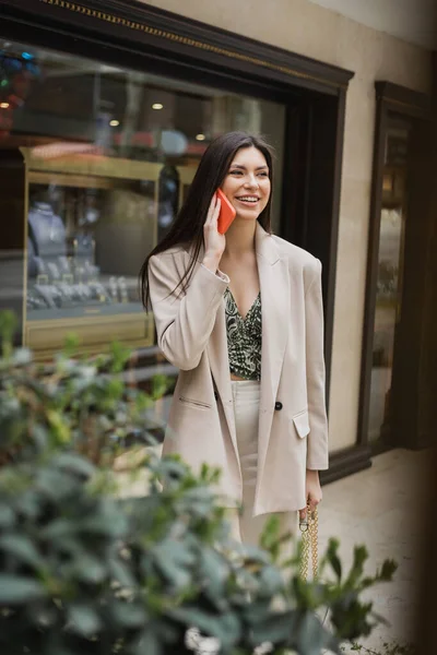 Smiling young woman with brunette long hair and makeup talking on smartphone and standing in trendy outfit with handbag on chain strap near blurred jewelry store and plant in Istanbul — Stock Photo